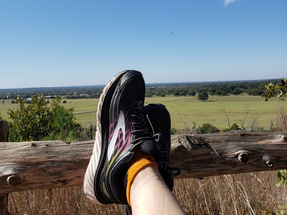 Feet in running shoes propped up against a wooden post fence in front of a scenic overlook of a green meadow