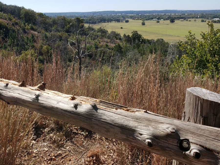 A wooden fence in front of a scenic view overlooking a tree-covered hill and a meadow.